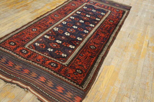 Low-pile red and blue rug with floral and geometric patterns sits diagonally on light, worn wood floors.