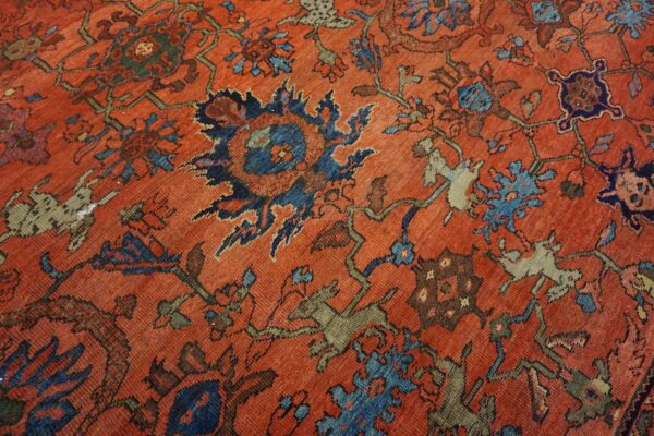 Worn, red-orange rug features stylized blue flowers, geometric forms, and khaki animal figures.