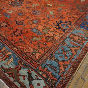 A rust orange rug with a dense floral pattern and contrasting blue borders rests on distressed wood flooring.