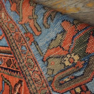 A tightly woven rug with red and light blue geometric designs is curled on distressed wood.