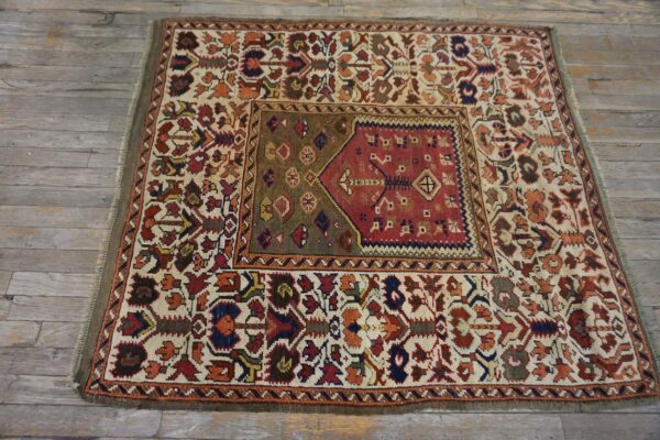 Small traditional rug with red, olive, and ivory geometric patterns lying on distressed gray wood flooring.