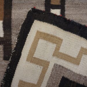 A corner of a flatweave rug is folded, displaying stepped geometric patterns in cream, tan, dark brown, and taupe.