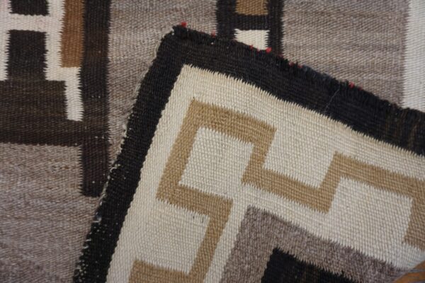 A corner of a flatweave rug is folded, displaying stepped geometric patterns in cream, tan, dark brown, and taupe.