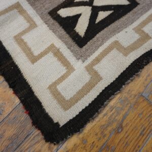 Low-pile geometric rug corner in cream, tan, and dark brown resting on distressed wood flooring.