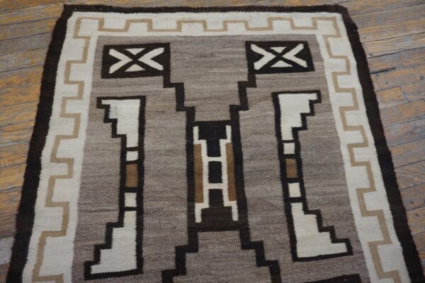 Flatweave rug featuring a stepped geometric design in brown, white, tan, and gray on distressed wood flooring.