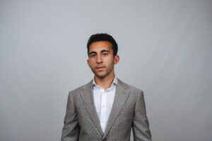 A man wearing a light gray suit and white shirt stands against a seamless light gray background.