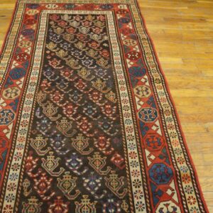 Dark runner rug with multicolored geometric patterns and red border sits on worn, light wood flooring.