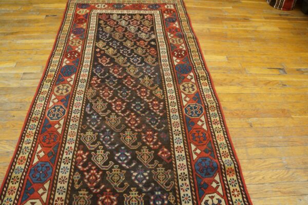 Dark runner rug with multicolored geometric patterns and red border sits on worn, light wood flooring.