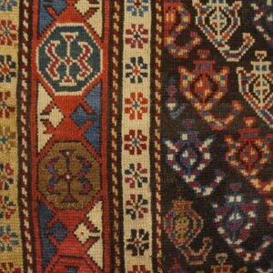Detailed view of a low-pile rug with geometric patterns in dark, red, blue, and tan on wood floors.