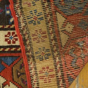 Knotted rug edge and flat woven backing in red, blue, and gold resting on distressed wood floor.