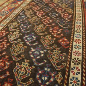 Low-pile rug with dark field, geometric red and blue motifs, and intricate borders rests on wood floor.