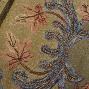 Ornate high-pile rug with pink leaves and blue paisley scrolls on a speckled khaki background next to wood flooring.