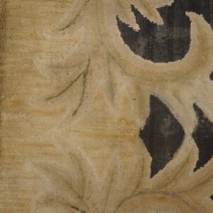 Detail of a rug showing light beige scrolling elements on a dark, pebbled texture framed by wood.