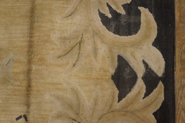 Detail of a rug showing light beige scrolling elements on a dark, pebbled texture framed by wood.
