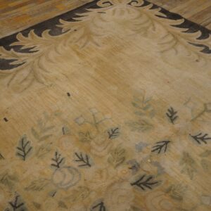 Close view of a tan rug with a dark scroll border and scattered blue and green floral patterns on wood planks.
