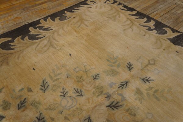 Close view of a tan rug with a dark scroll border and scattered blue and green floral patterns on wood planks.