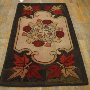 Dark floral hooked rug with red and tan leaves on a cream center, set on worn wooden floorboards.