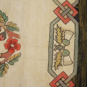 A textured loop rug displays a red floral wreath and geometric border against a cream field on wood flooring.