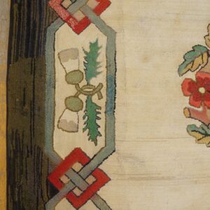 Cream hooked rug detail featuring red flowers, green foliage, and a multicolored geometric border on distressed wood.