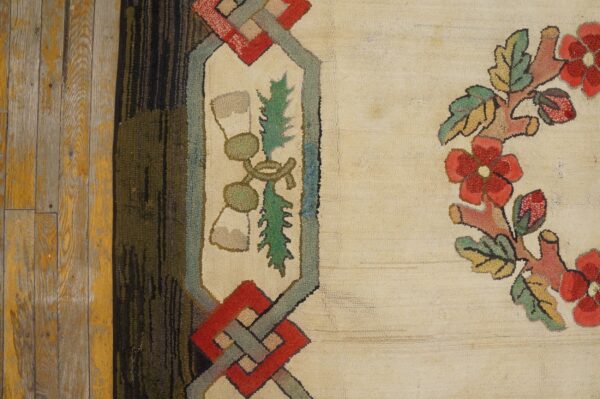 Cream hooked rug detail featuring red flowers, green foliage, and a multicolored geometric border on distressed wood.