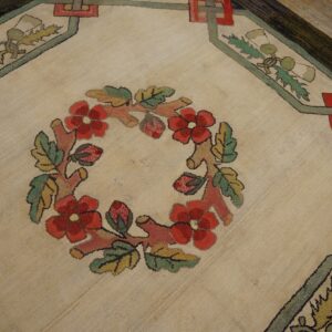 Textured, low-pile rug showing a central red floral wreath and geometric borders on wood planks.
