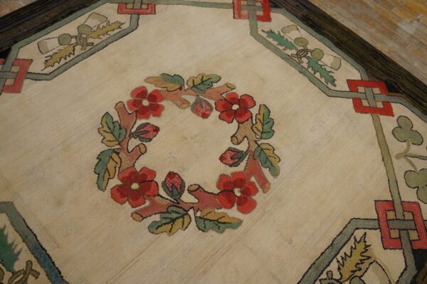 Textured, low-pile rug showing a central red floral wreath and geometric borders on wood planks.