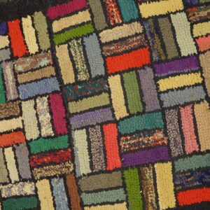 Colorful hooked rug with a rectangular block pattern and black borders rests on light wood planks.