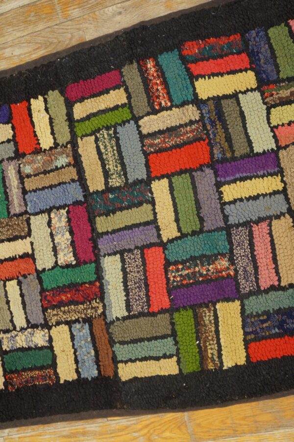 Colorful hooked rug with a rectangular block pattern and black borders rests on light wood planks.