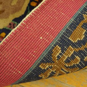 A folded rug shows a pink woven backing and dark blue, brown, and tan pile against a yellow wood floor.