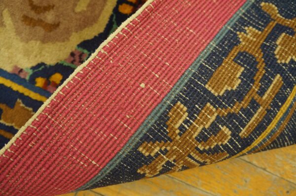 A folded rug shows a pink woven backing and dark blue, brown, and tan pile against a yellow wood floor.