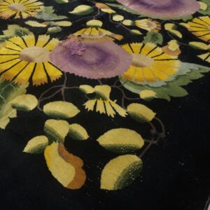 Black rug with high pile, patterned with bold yellow, green, and purple flowers on wood flooring.