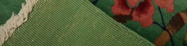 View of the rug reverse showing ribbed light green weaving and fringe next to the deep pile green, red, and brown pattern.