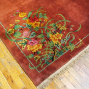 Close-up of a high-pile rust rug with bright floral and vine designs on worn yellow wood planks.