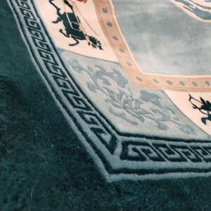 Teal and cream rug detail showing carved high pile, geometric border, and stylized figures on the edge.
