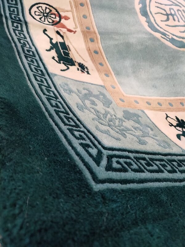 Teal and cream rug detail showing carved high pile, geometric border, and stylized figures on the edge.