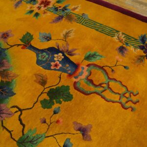 Yellow-gold rug with stylized flowers and vines, a blue vase motif, and a visible burgundy edge on wood flooring.