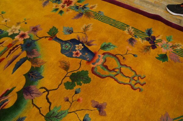 Yellow-gold rug with stylized flowers and vines, a blue vase motif, and a visible burgundy edge on wood flooring.