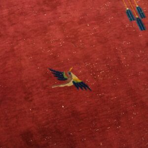 Detail of a red medium-pile rug showing a stylized flying bird and blue hanging tassels.