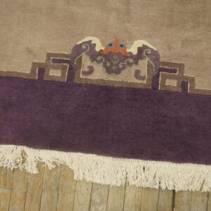 Beige rug with deep purple border features a multicolored crest detail above white fringe on wood planks.