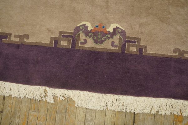 Beige rug with deep purple border features a multicolored crest detail above white fringe on wood planks.