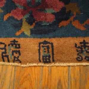 Teal, red, and gold floral rug with a brown inscription border lies on a distressed wood floor.