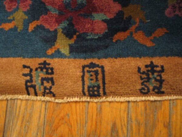 Teal, red, and gold floral rug with a brown inscription border lies on a distressed wood floor.
