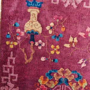 Close-up of a magenta rug featuring multicolored floral and vase motifs, bordered by a light pink fret pattern.