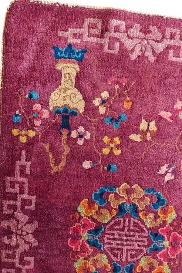 Close-up of a magenta rug featuring multicolored floral and vase motifs, bordered by a light pink fret pattern.
