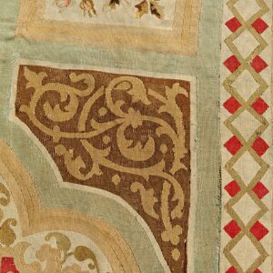 Detailed rug view featuring brown scrollwork, sage green panels, and a red diamond geometric border.