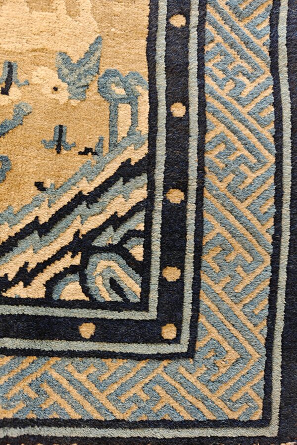 Close-up of a tan, slate blue, and dark blue rug showing geometric and floral border patterns with a medium pile.