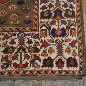 Ornate tribal rug corner on wood floors, showing an olive field and a cream border with red, blue, and orange motifs.
