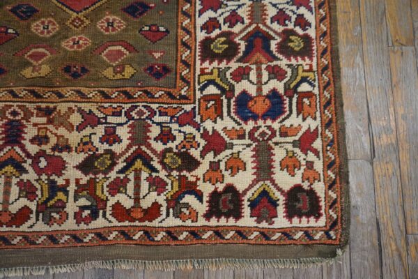 Ornate tribal rug corner on wood floors, showing an olive field and a cream border with red, blue, and orange motifs.