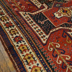 Geometric red and dark blue rug showing borders with stylized figures rests on aged wood plank flooring.