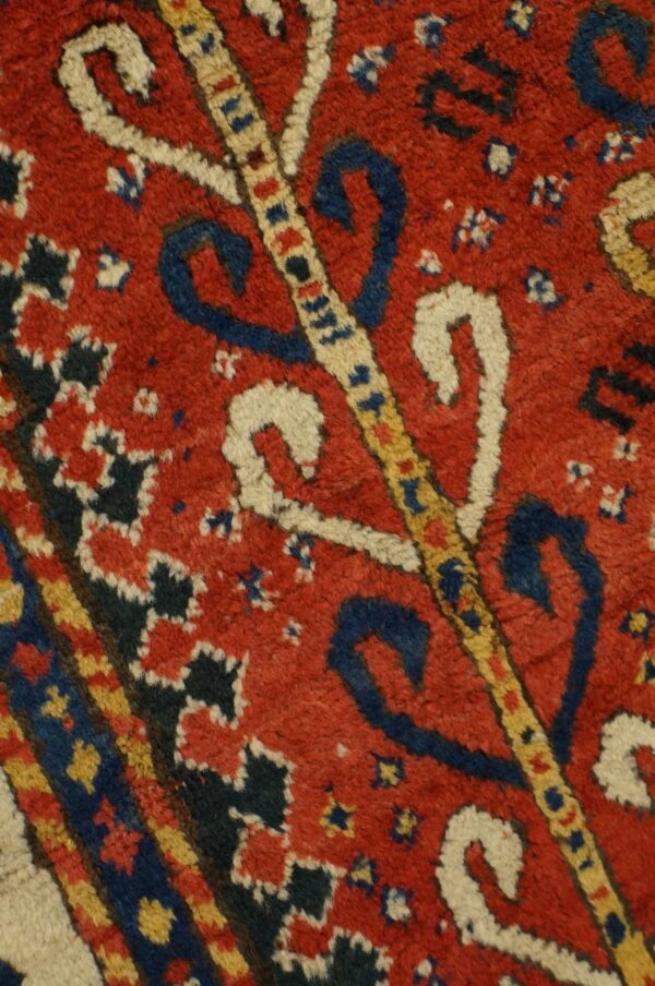 A close-up of a textured rug showing a red field with dark blue, cream, and yellow scrolling motifs.